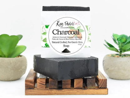 Activated Charcoal black rectangular bar soap with white and green natural crafted label resting on wooden soap dish with plants behind