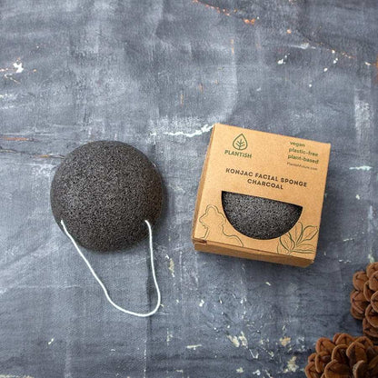 Konjac Facial Sponge in charcoal grey, round porous texture with a white hanging string, eco-friendly packaging box nearby