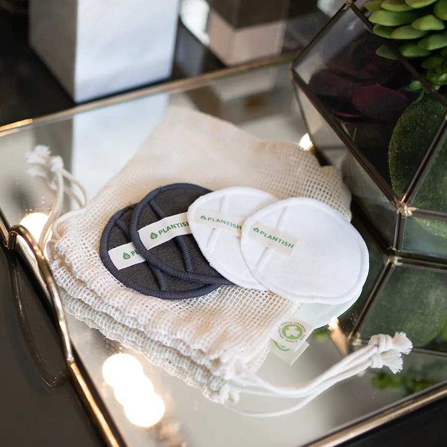 Reusable Cotton Rounds in white and charcoal with a soft texture, displayed alongside a mesh storage bag on a reflective surface.